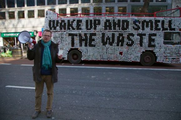A man with a foghorn and a cup standing in front of a double decker bus with coffee cups on its side that spell out 'Wake up and smell the waste.'