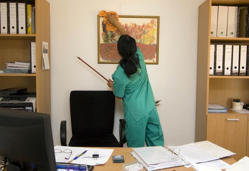 A dark-haired woman wearing a green uniform cleaning an office, specifically dusting a picture on the wall.