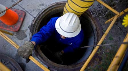 A worker in a blue uniform and a white hard hard climbing out of a manhole.