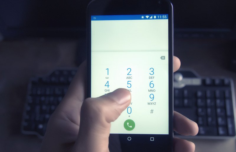 A person holding a black phone which is on the dialing screen with a keyboard in the background.