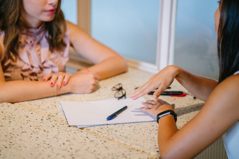 Two women, one in a pink top with pink nail varnish and lipstick on, and the other dark-haired with a black digital watch on her wrist, talk sat at a table next to a window with paper, a pair of glasses and pens in the middle.
