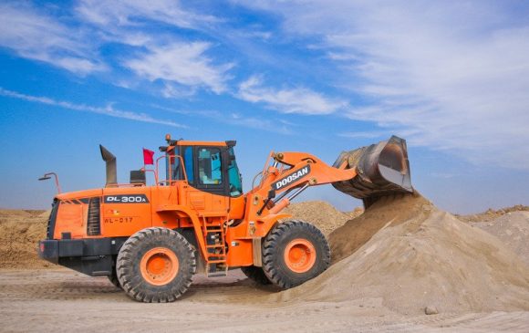 An orange and black excavator picking up sand against a blue sky with some clouds in it.