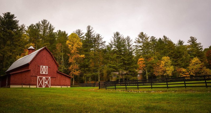 A red farm in a field with a fence and trees around it.