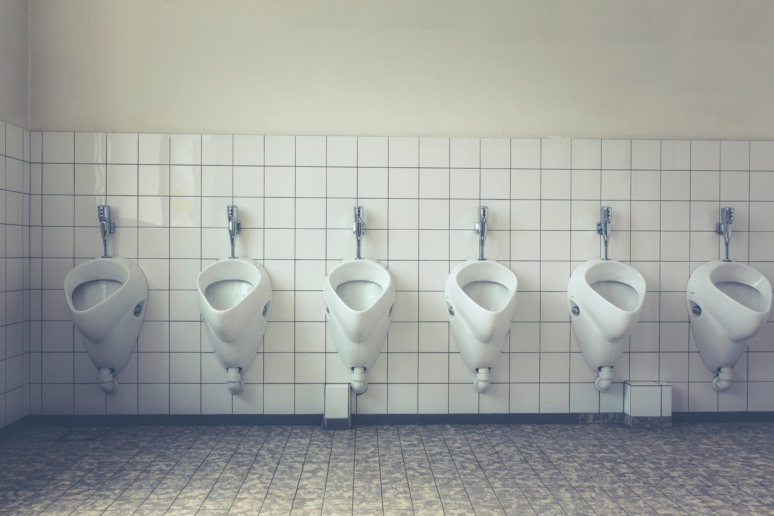 A line of urinals against a white tiled wall in a public bathroom.