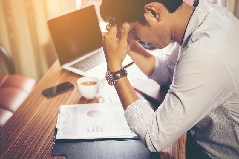 A man sat at a desk which has a laptop, papers and a phone on it. He is holding his heads in his hands, looking stressed.