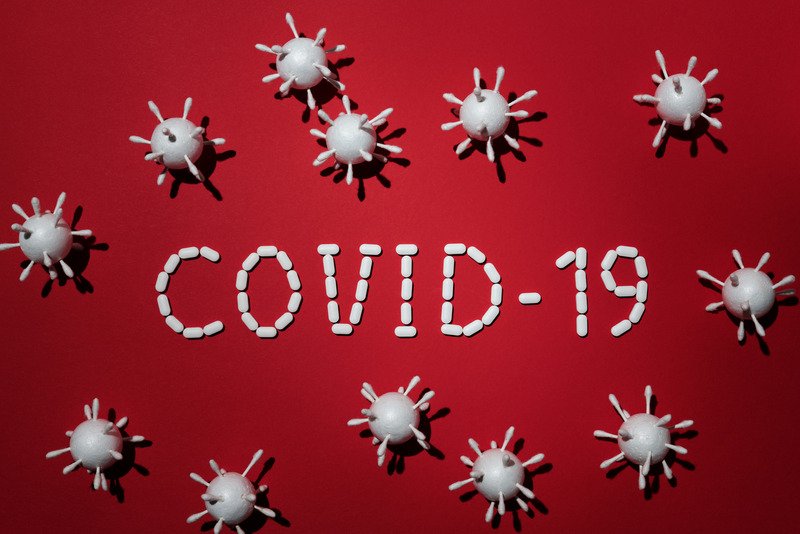 COVID-19 spelt out in small white tablets surrounded by microscopic viruses against a red background.