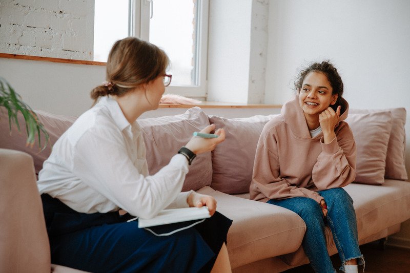 A young women talking with a therapist whilst both are sat on a pink sofa facing towards each other.