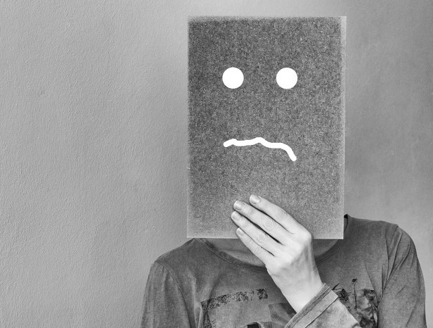 A black of white picture of a person covering their face with a sheet with a sad face drawn on it.