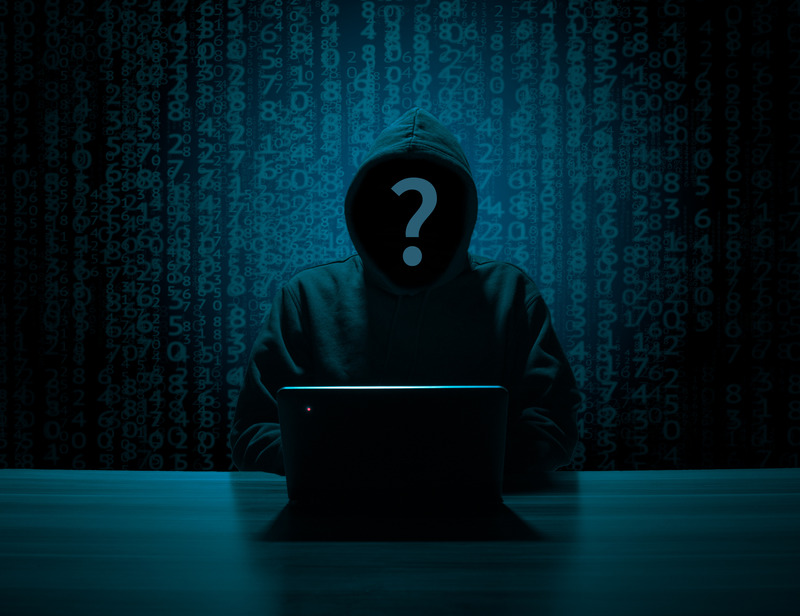 An unknown person with a hood up and a question mark where their face should be sitting in front of a laptop on a table. The background is strings of coding.