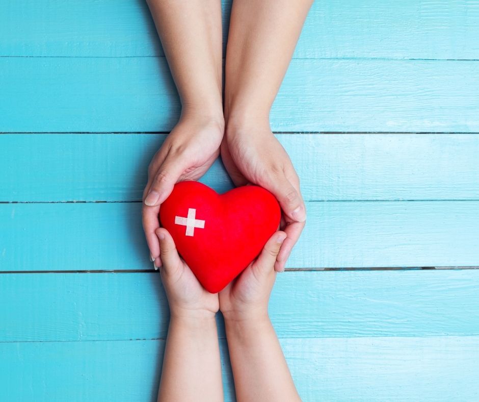 Two sets of hands holding a heart (cartoon shape) with a white cross on it