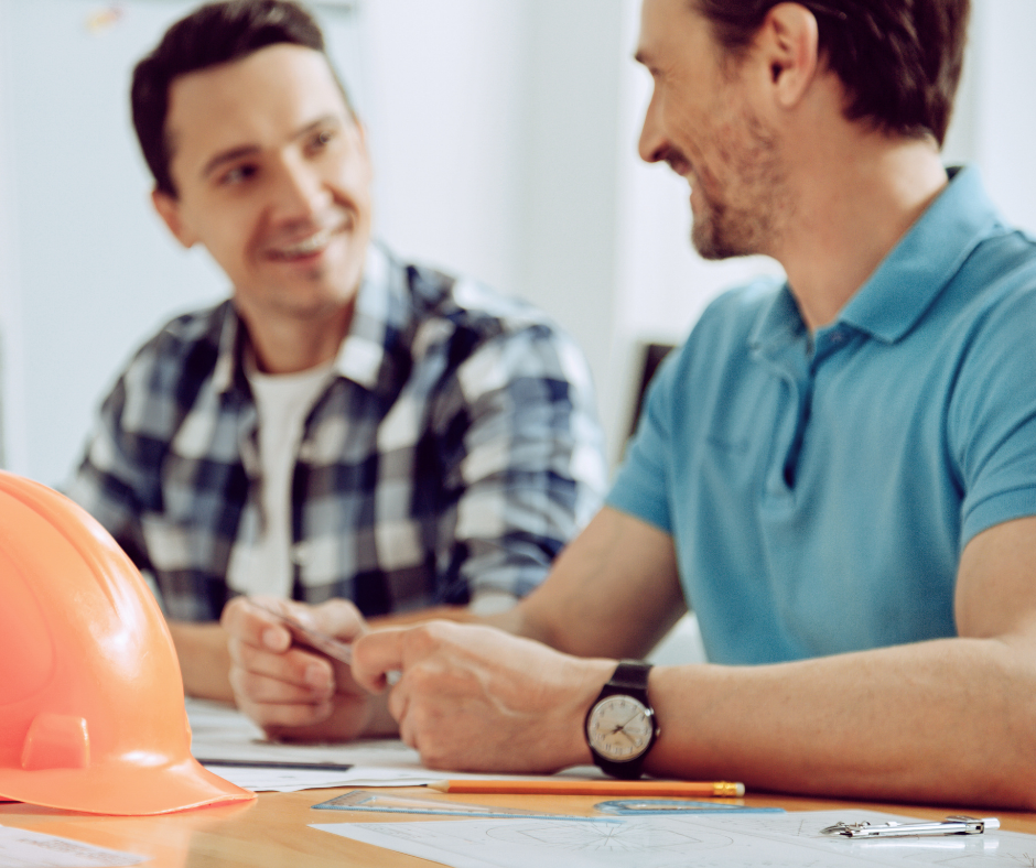 Two men talking at a desk, which has papers and a construction helmet on it
